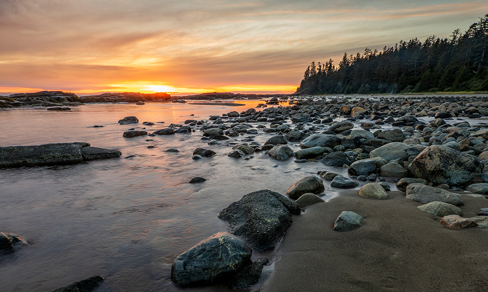 West Coast Trail: Sunset in front of the water, with big rocks in it and a part of the beach showing